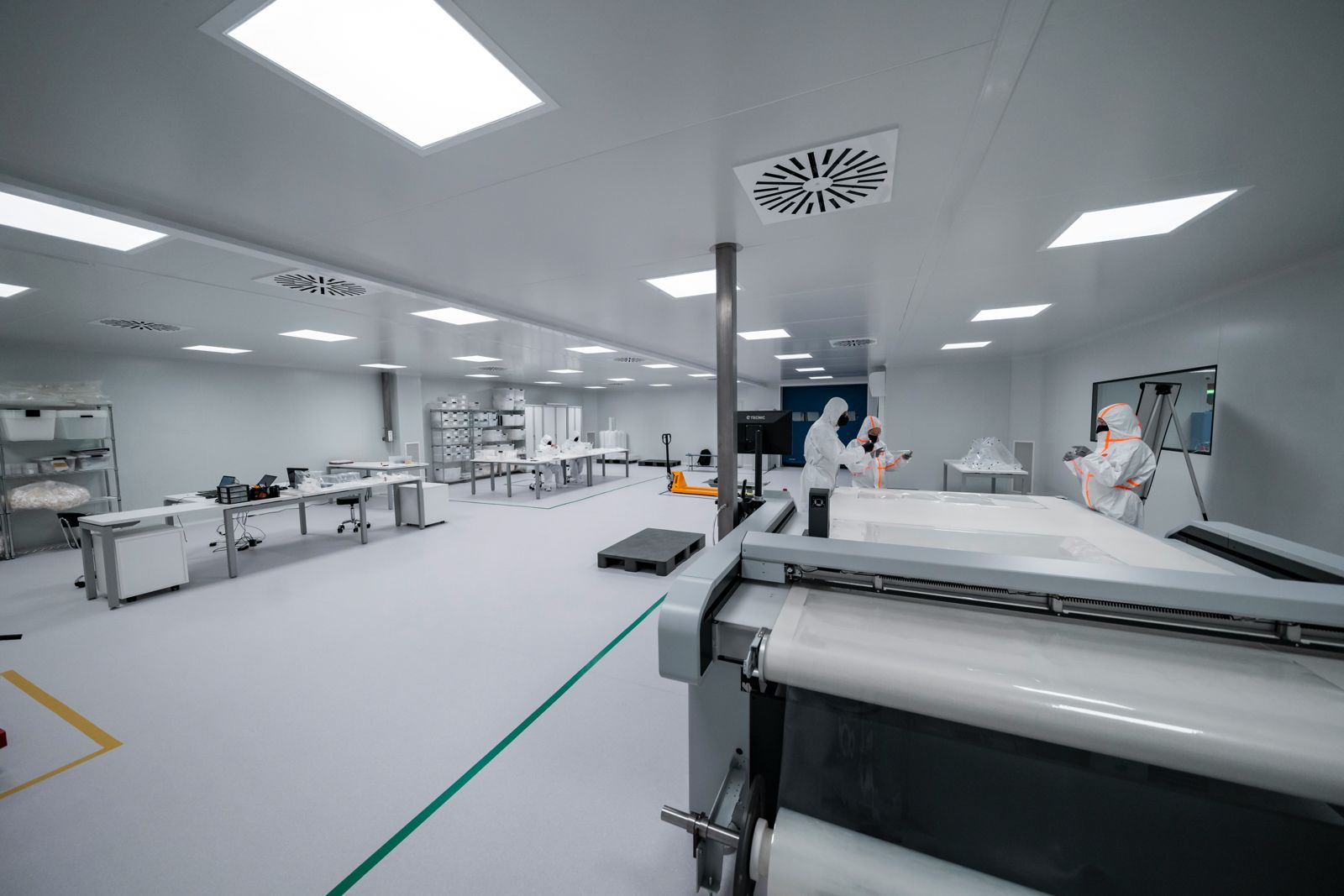 Bright cleanroom production floor with operators in hooded white coveralls at a fabric cutting table