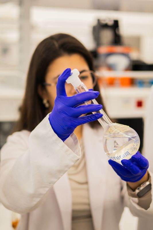 Lab worker in a white lab coat and purple nitrile gloves holding a volumetric flask