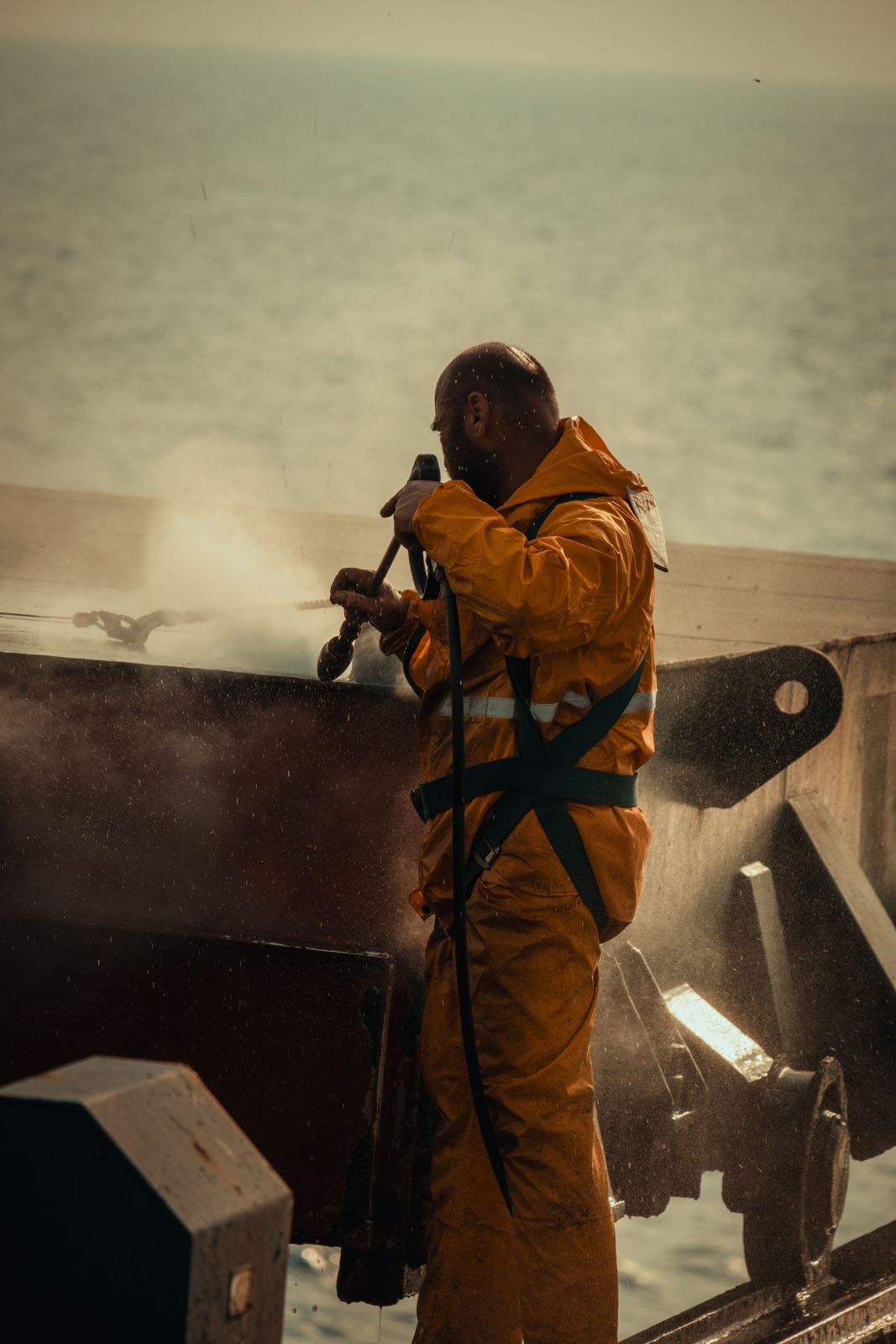 Worker in an orange PVC rain suit pressure-washing a ship deck at sea