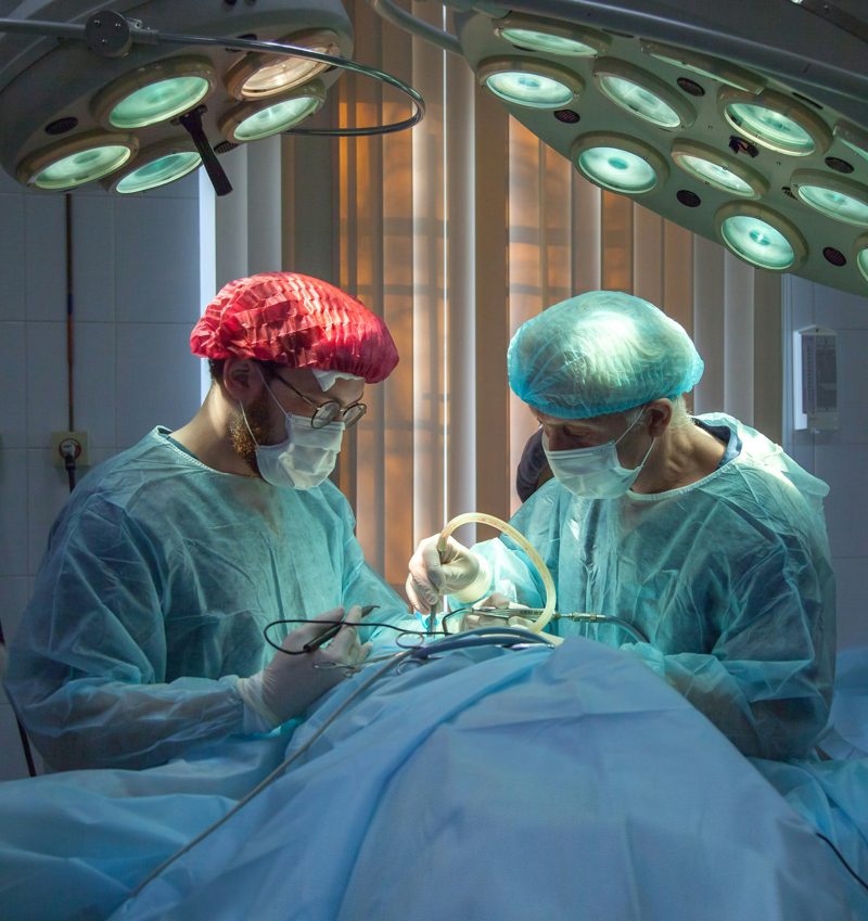 Two surgeons in disposable gowns, bouffant caps and masks under operating lights